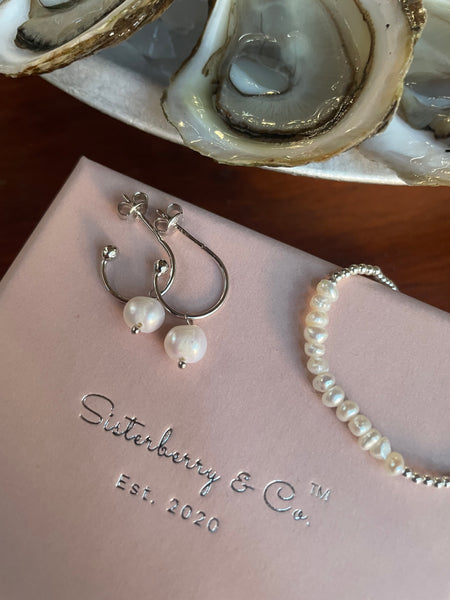 Silver hoop earrings with white freshwater pearls and a matching pearl bracelet, displayed on a pink Silterberry & Co. box next to fresh oysters.