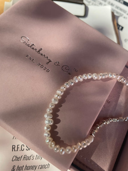 A strand of white freshwater pearls draped over a dusty pink jewelry box printed with the brand name 'Sisterberry & Co'.