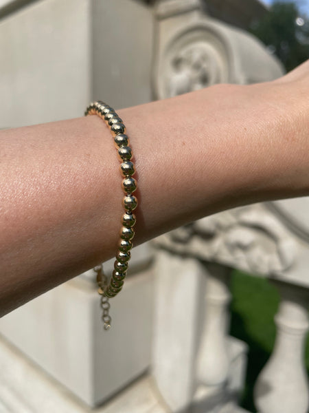 Woman wearing gold beaded ball bracelet on wrist/arm with a beige architecture in the background