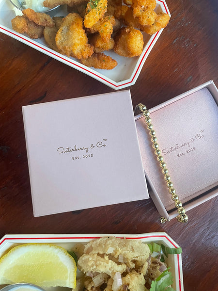 Blush pink Sisterberry & Co box with 6mm round ball beaded bracelet with fried calamari and clams in the background. 