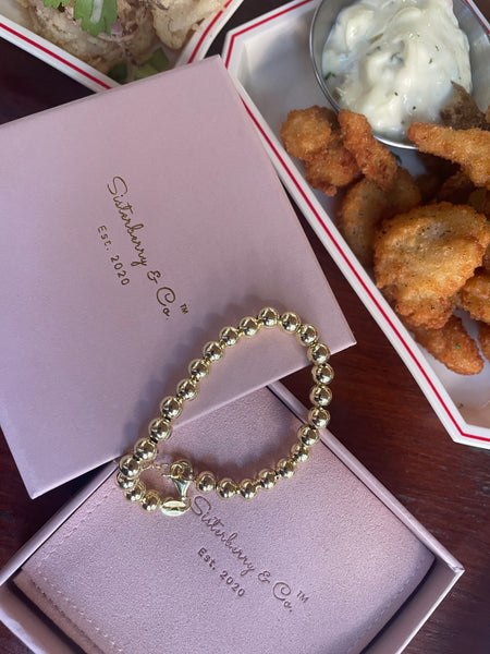 A gold beaded bracelet with a small charm, presented on a light pink Sisterberry & Co. box next to a tray of fried food and a dipping sauce.