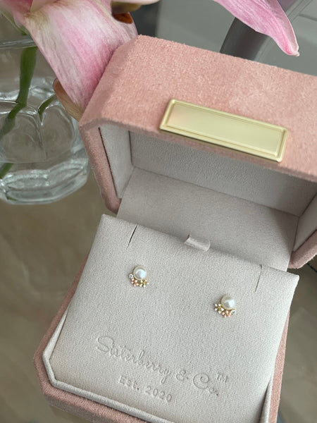 A pair of gold and pearl stud earrings displayed on a light pink velvet jewelry box by Sisterberry & Co, with a pink flower in the background.