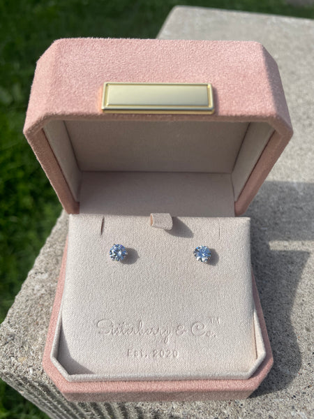 A pair of small round stud earrings with clear gemstones displayed in an open pink jewelry box on a stone surface outdoors.