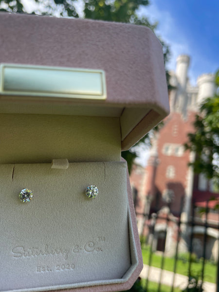 A pair of round-cut stud earrings displayed on a velvet jewelry box from Satinsky & Co., photographed outdoors with a blurred brick building and greenery in the background.