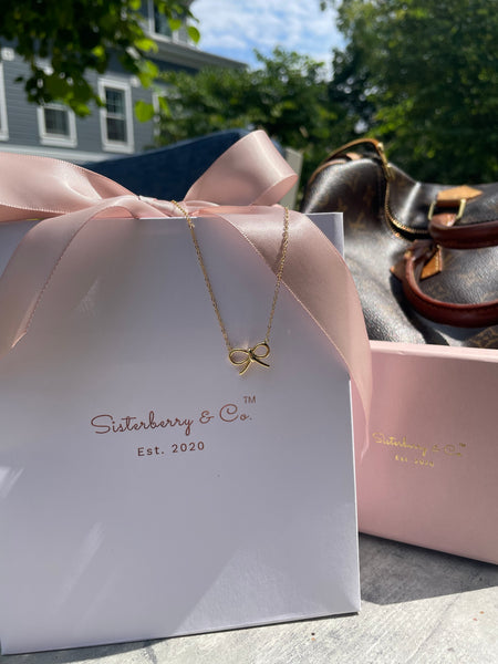 A gold bow pendant necklace from Sisterberry & Co. is displayed on its white gift box with a pink satin ribbon, with a brown leather handbag and a pink presentation box in the background outdoors.