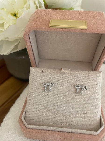 A pair of silver bow stud earrings with clear crystals, displayed on a branded pink velvet jewelry box by Sutterberry & Co.