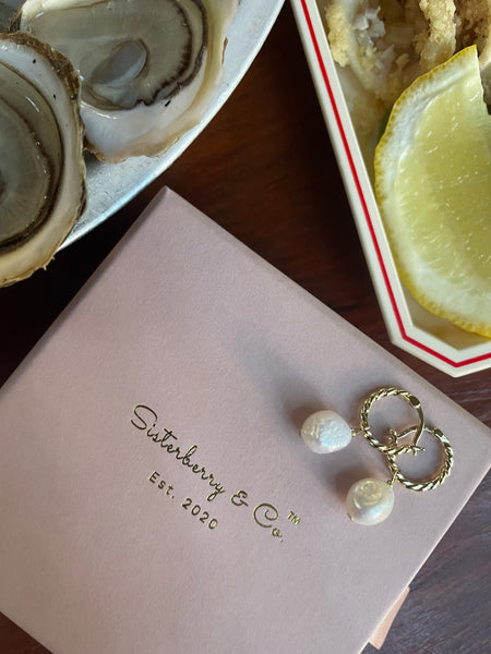A close-up of a pale pink jewelry box with gold foil branding reading 'Sisterberry & Co Est. 2020,' styled on a dark wooden table with oysters, lemon, and a gold twisted hipe earrings with pearls nearby.