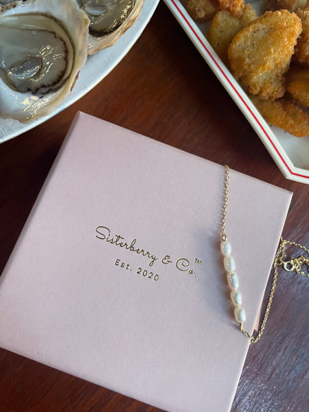 A delicate gold chain necklace with a vertical strand of white freshwater pearls, displayed on a pale pink Sisterberry & Co. gift box on a dark wooden table, with oysters and chicken nuggets in the background.