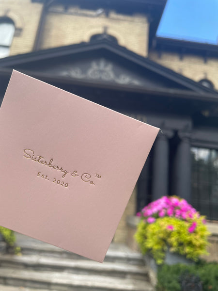 A square blush pink card or envelope with the brand name 'Sisterberry & Co Est. 2020' printed in gold foil, held in front of a blurred brick Victorian house and flowerbed.