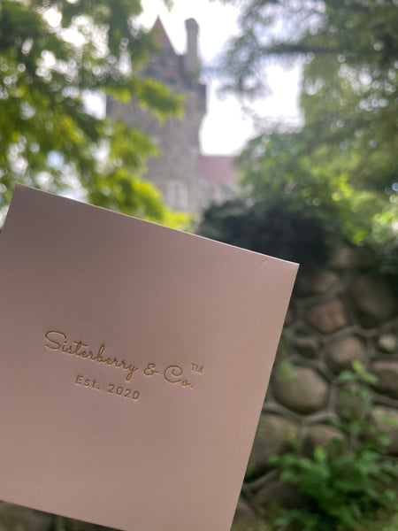A close-up of a pale pink card or envelope printed with the brand name 'Sisterberry & Co' and 'Est. 2020' in gold foil, held in front of a blurred outdoor background featuring green foliage and a stone wall with a castle-like building behind it.