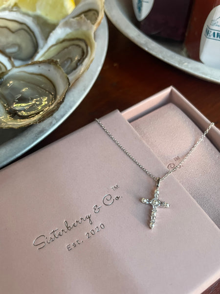 A silver cross pendant necklace with clear stones, displayed on a pink jewelry box with the brand name Sisterberry & Co. visible, with oysters in the background.
