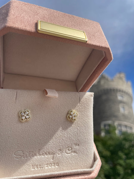 A pair of gold stud earrings with clear gemstones displayed on a pink velvet jewelry box by Sisterberry & Co, set against a blurred outdoor background of a stone tower and blue sky.