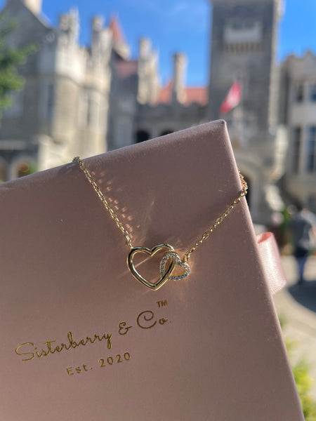 A gold double-heart pendant necklace displayed on a pink Sisterberry & Co gift box, with a blurred castle background and blue sky.