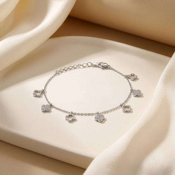 A delicate silver chain bracelet with five small flower charms, each adorned with clear stones, displayed on a cream-colored tray with draped satin fabric.
