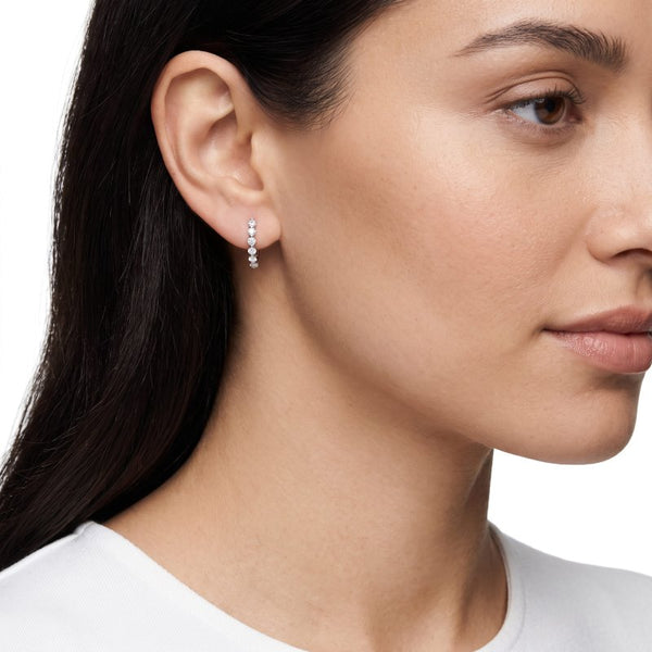 Woman with dark black hair wearing lab diamond huggie earrings in sterling silver rhodium plated with a white tshirt.