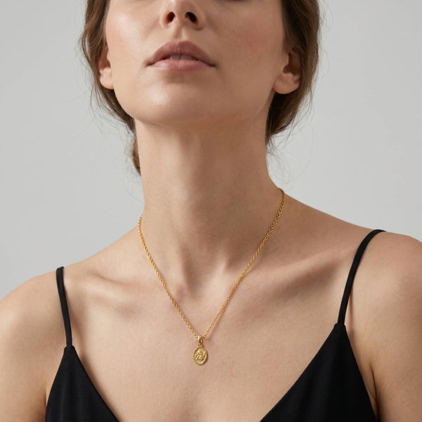 A close-up of a model wearing a delicate gold chain necklace with a small circular pendant, styled with a black spaghetti-strap top against a neutral gray background.