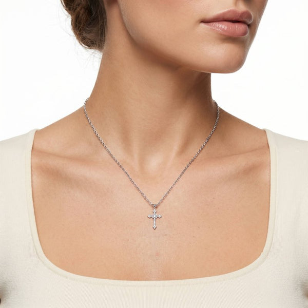 A close-up of a model wearing a delicate silver cross pendant necklace on a fine chain, paired with a cream-colored scoop-neck top against a white background.