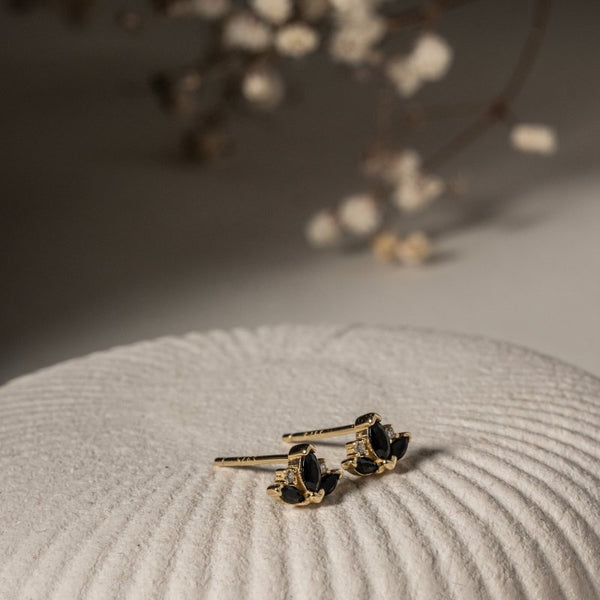 Gold earrings with black stones on a textured surface with blurred flowers in the background